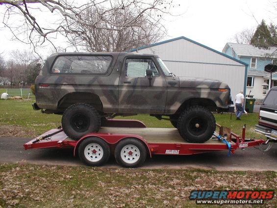 Camouflaged Ford Bronco on a red trailer, side view. Another vehicle is partially visible on the right