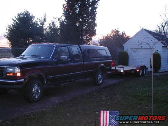 Black and maroon Ford F-Series truck with a camper shell, towing a flatbed trailer. Parked in a driveway next to a white garage