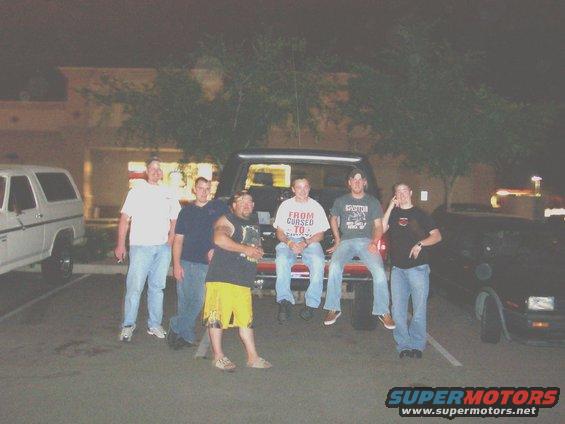 group of guys standing around and sitting on the tailgate of a truck, with a white SUV and a dark car nearby