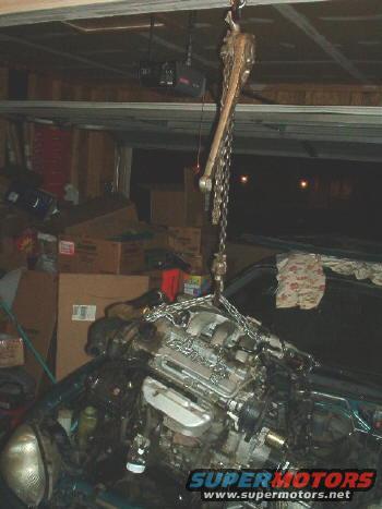 Engine suspended by a chain hoist in a garage, with boxes in the background