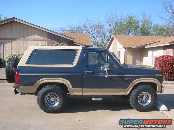 Side view of a blue 1986 Ford Bronco with a tan top and spare tire on the back
