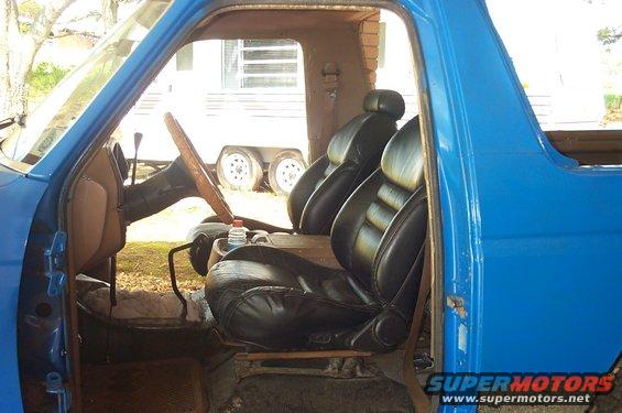 Interior view of a blue truck showing black bucket seats and a wooden steering wheel. A trailer is visible through the open door