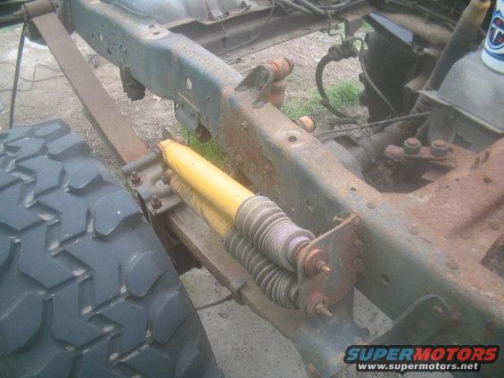 Close-up of a rusty truck frame showing a leaf spring and a yellow shock absorber. A large tire is visible on the left