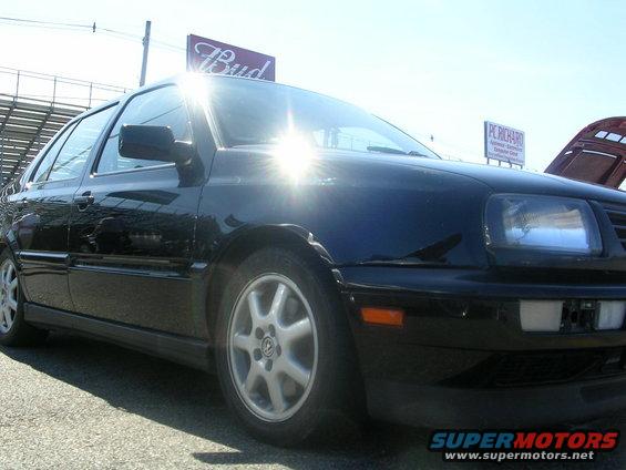 Black Volkswagen sedan, front quarter view, with a "Bud" sign and stadium seating in background