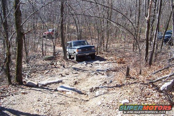 Two off-road vehicles navigating a rocky, wooded trail. One blue SUV is in the foreground, with a red truck and a dark SUV further back