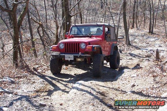 Red Jeep Wrangler with off-road tires on a rocky, wooded trail