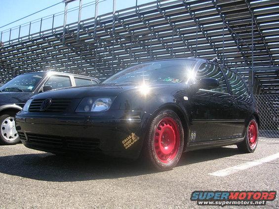 Black Volkswagen Jetta with red steel wheels parked on asphalt, stadium seating in background