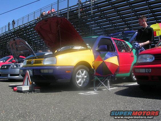 Multi-colored Volkswagen Golf with hoods open, parked in front of bleachers