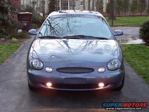 Front view of a gray sedan parked in a driveway. The car has oval headlights and a mesh grille
