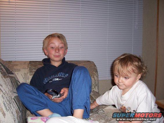 Boy in Seahawks hoodie holds flip phone while toddler plays on couch