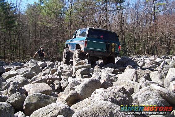 Green Ford Bronco with large tires navigating a rocky trail. A person stands in the background
