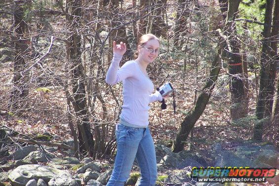 Woman with glasses and a light pink shirt holds a camcorder while walking in a wooded area with bare trees and rocks