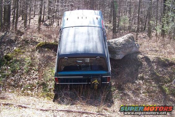 Back view of a dark green 1979 Ford Bronco wedged against a large rock on a wooded trail