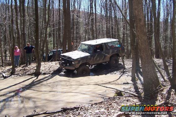 Muddy 1978 Ford Bronco navigating a water obstacle in a forest, another SUV behind it