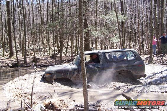 Dark colored Ford Bronco driving through a muddy creek in a wooded area. Some people are watching from the bank