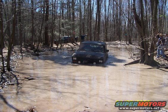 Two off-road vehicles navigate a muddy water crossing in a dense forest