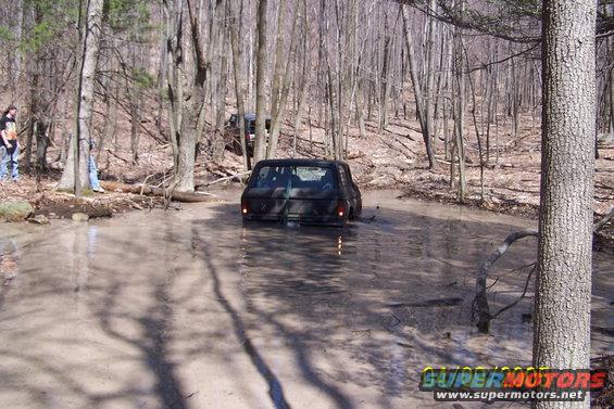 Black SUV driving through muddy water in a forest, rear view