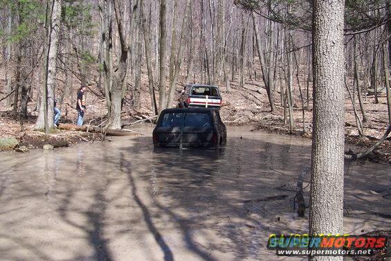 Black SUV stuck in mud, rear view. Another SUV behind it in a wooded area