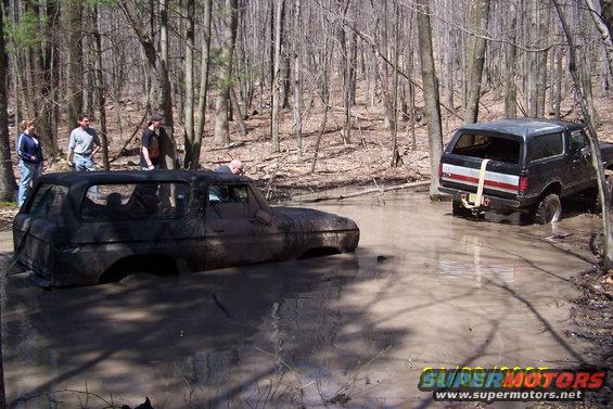 Two SUVs stuck in mud in a wooded area. One is a dark color, the other is black with a red and white stripe on the rear. People stand nearby