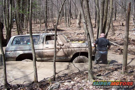Muddy 1978 Ford Bronco stuck in a wooded area, with a person nearby