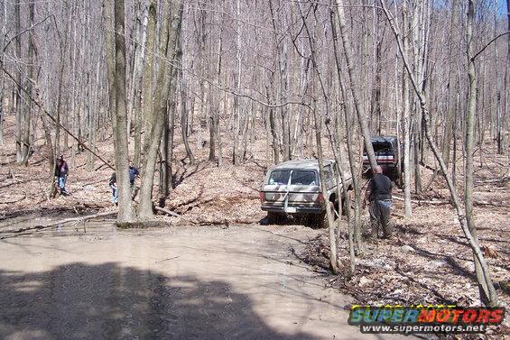 Tan 1978 Ford Bronco driving on a muddy forest trail, rear view