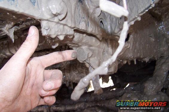 Hand pointing at a bolt on a muddy vehicle's undercarriage