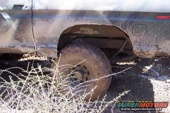 1979 Ford Bronco rear wheel well and tire covered in mud, with dry brush in foreground