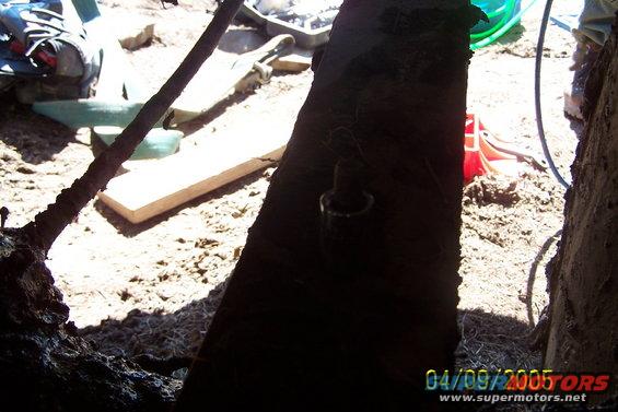 Dark, rough metal frame of a vehicle chassis in dirt, with a wooden plank and green hose visible
