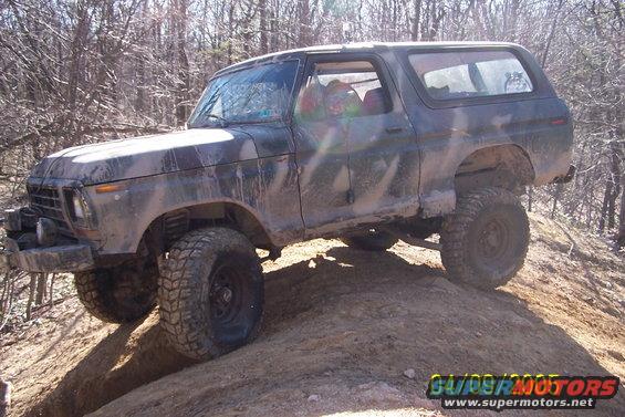 Muddy Ford Bronco climbing a dirt hill, trees in background