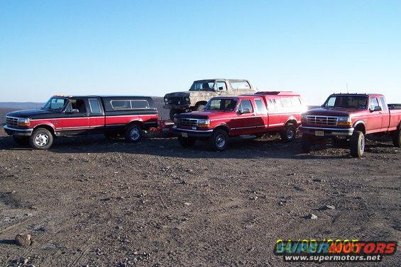 Four trucks and a Bronco parked on a dirt lot under a clear sky