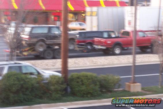 Black Ford Bronco on a trailer being towed by a red truck. Another truck is behind them