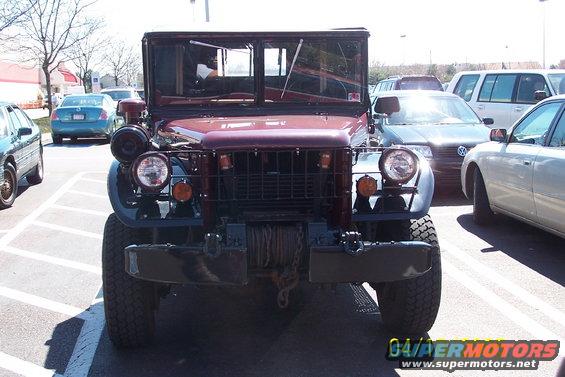Front view of a maroon military truck with a winch. Other cars are parked nearby
