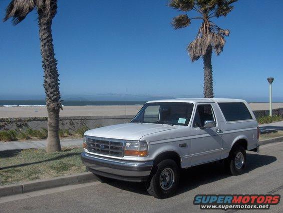 White 1992 Ford Bronco parked on a street next to a beach, with palm trees and ocean in the background. The vehicle is seen from the front-right