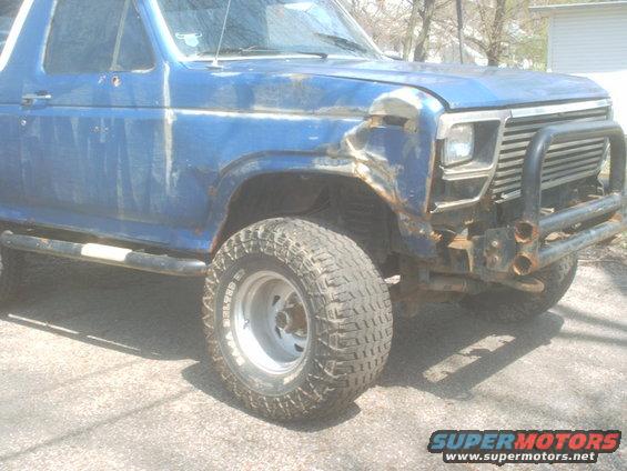 Blue Ford Bronco with a dented fender and aftermarket bumper. It has large off-road tires