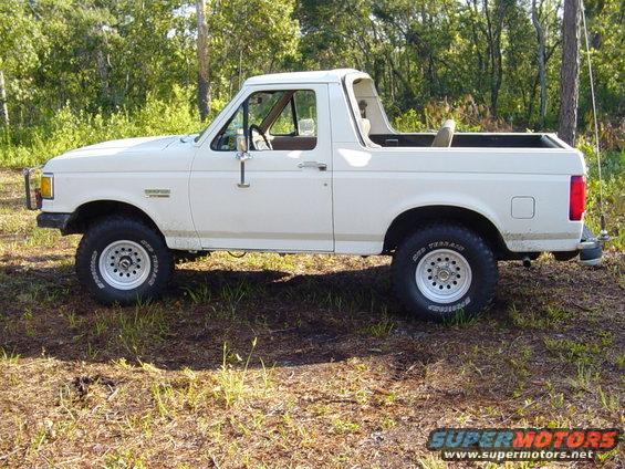 White Ford Bronco parked on grassy ground with trees in background. Side view