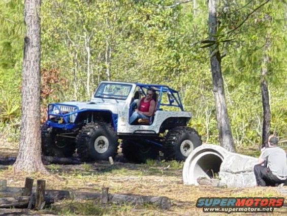 White and blue off-road jeep with large tires and a roll cage, driven by a woman