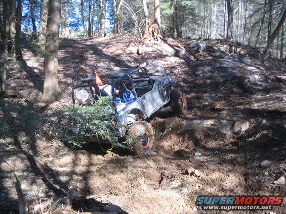 Off-road buggy with roll cage navigating a muddy, wooded trail. Orange flag on antenna