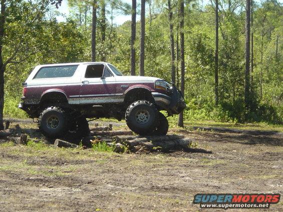 1993 Ford Bronco with large tires driving over logs in a wooded area