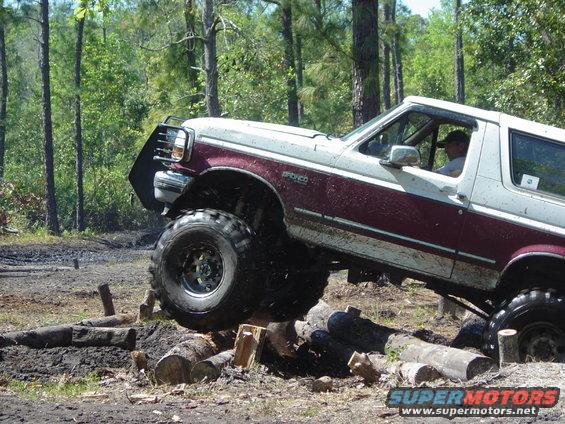 Two-tone maroon and white Ford Bronco with lifted suspension and large tires navigating an obstacle course of logs