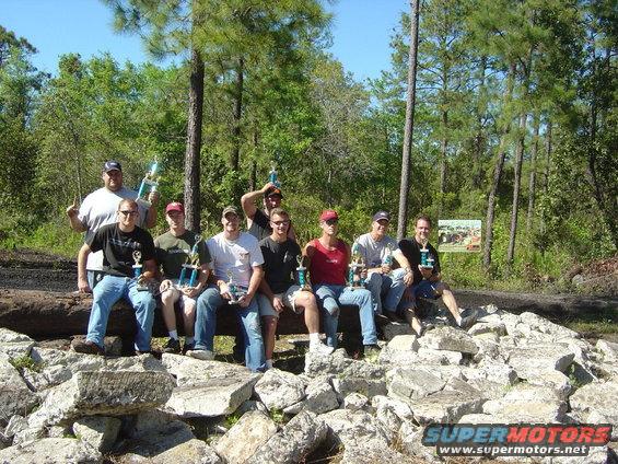 Group of men sitting on a log and rocks, holding trophies. Trees and sky in background
