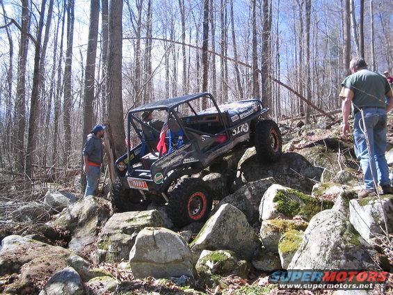off-road buggy with roll cage climbing over large rocks in a wooded area, people watching