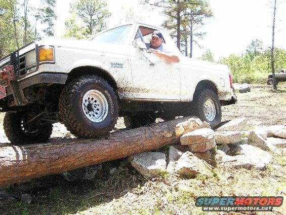 White pickup truck driving over a log obstacle. Another truck is visible in the background