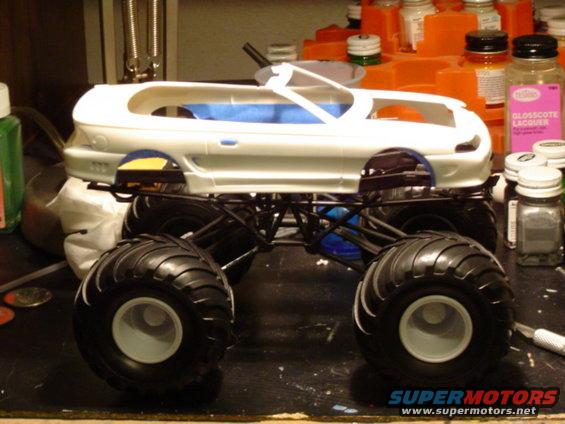 White model monster truck body on a black chassis with large tires, on a workbench with craft supplies