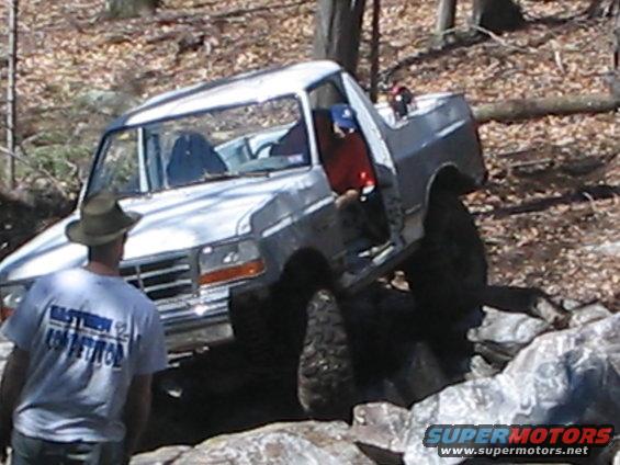 White Ford F-150 truck on rocks, driver's door open, man in hat watches