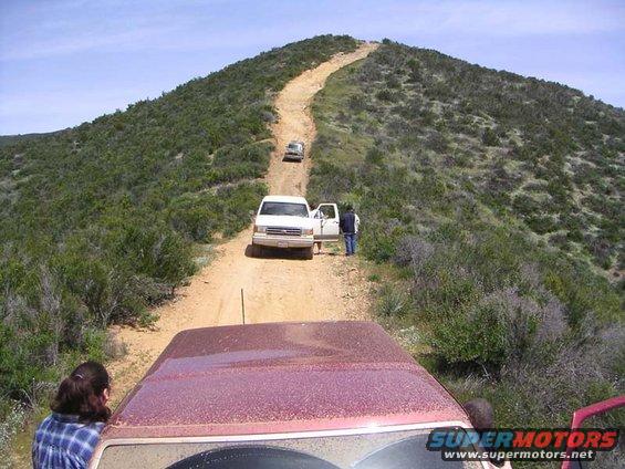 Red Ford Bronco hood and windshield in foreground, white Ford Bronco on dirt road ahead