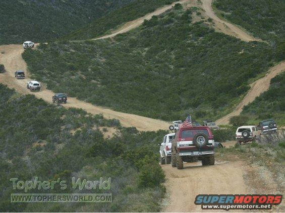 Red Ford Bronco with American flag on antenna drives on dirt road behind other SUVs