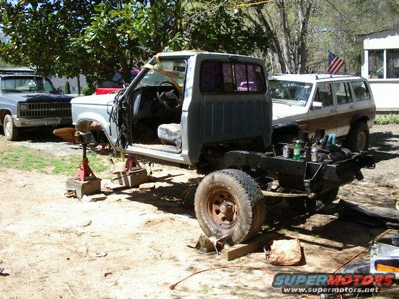 1985 Ford Bronco chassis on jack stands with front end removed, another truck and SUV in background