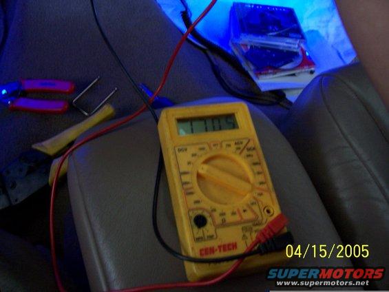 Interior view of a vehicle with a yellow Cen-Tech multimeter connected to wires, next to pliers and a pry tool. CDs are stacked in the background