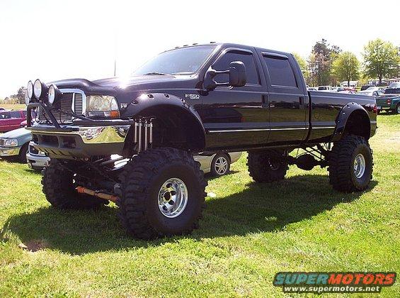 Lifted black pickup truck with big tires and extra lights on the front. Other vehicles are parked in the background
