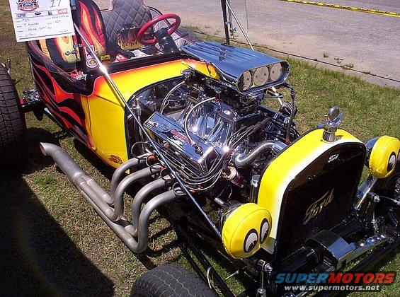 Custom hot rod with yellow and flame paint, exposed V8 engine, and large exhaust pipes. Ford logo on grille